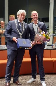 Sjoerd Geissler (rechts), met lintje opgespeld en bloemen, poseert met Burgemeester Oskam (links), met ambtsketting om.